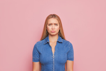 A woman in denim dress with sad expression in front of pink background