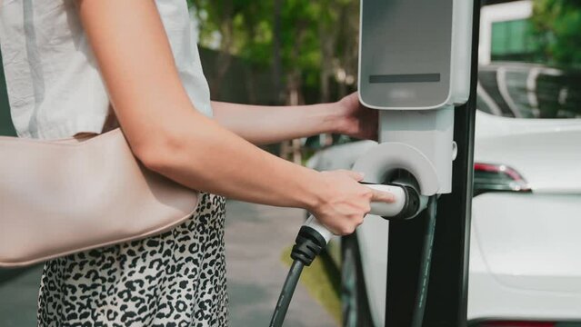 Slow motion woman recharge EV electric car battery at residential area EV charging station in urban city condo with sustainable green and renewable clean energy lifestyle for electric vehicle innards