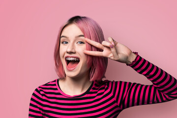 Photo image - happy pin haired beautiful woman in braces show v-sign victory peace hand gesture two fingers, with opened mouth, isolated wall background. Student girl at studio. Dental care ad concept