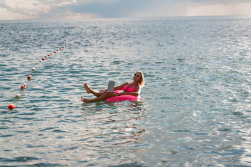 Woman works on laptop in sea. Freelancer, blond woman in sunglases floating on an inflatable big pink donut with a laptop in the sea at sunset. People summer vacation rest lifestyle concept.