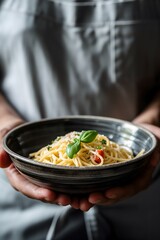 A person in a grey apron is holding a bowl of pasta, spaghetti