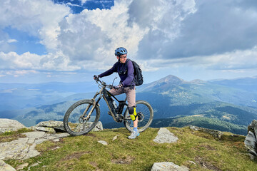 Fototapeta premium Cyclist man riding electric bike outdoors. Male tourist resting on the top of hill, enjoying beautiful mountain landscape, wearing helmet and backpack. Concept of active leisure.