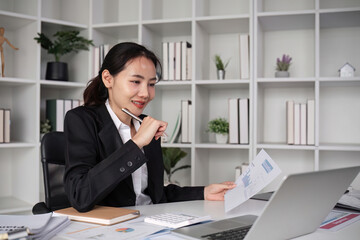 Young Asian business woman in a suit sits and reviews company work documents, checking information on a laptop on a work desk in a white office.