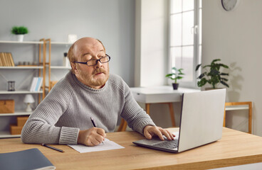 Senior man in eyeglasses using laptop and making notes on paper. Bearded elderly man wearing...