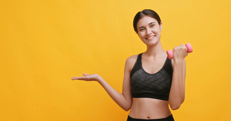 Young sportswoman shows off her physical strength by lifting dumbbell. Isolated on yellow background in studio.