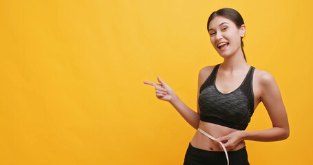 Young female in sportswear using a tape measure to measure her waist and pointing at something. Isolated on yellow background in studio.