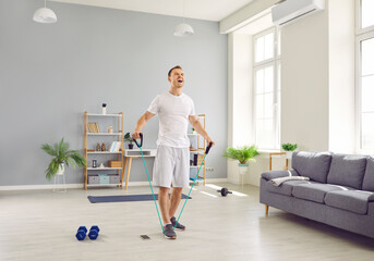 Young purposeful man at home doing sports strength training using rubber resistance band. Man in t-shirt and shorts shouting while stretching elastic band in living room. Home fitness training concept