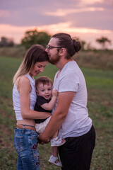 Fototapeta premium Diversity stylish father kisses mothers forehead while holding little son, concept of beautiful family connection against vibrant sunset. Family ties, togetherness. Close up portrait