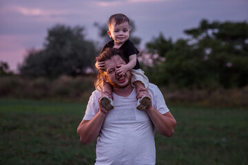 Obraz premium Diversity father with beard carrying son on shoulders shares tender moment in twilight lit field, embodying the essence of paternal love. Man and boy fooling around, playing. Child touches mans nose