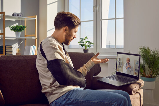Man with physical injury consulting online doctor on telemedicine clinic website. Young man with broken arm sitting on sofa with laptop computer and talking to doctor via video call