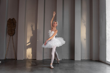 Poised young ballerina with feathered tiara, classic white tutu, stands on tiptoes in pointe, striking an elegant pose in brightly lit dance studio, shadow dancing on walls. Swan Lake ballet artist