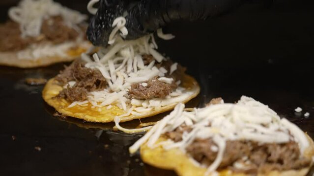 Shredded cheese added to birria tacos cooking on flat top grill, black gloved hand sprinkles grated quesadilla cheese to sizzling tacos, slow motion slider close up 4K