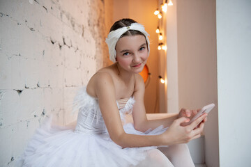 Close up portrait of young ballerina in white tutu, feather tiara, sits on the steps in loft...