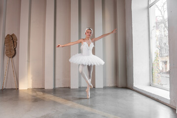 Fototapeta na wymiar Elegant young ballerina performs beside large windows, poised stance, delicate white tutu illuminated by natural light, embodying the fusion of strength, grace. White swan performance. Copy space