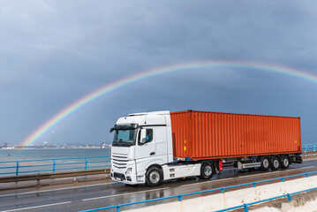 Truck with maritime container driving on a wet road and under a rainbow, side view.