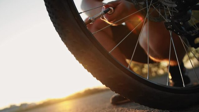 Bicyclist inflates rear flat tire by compact hand pump with retractable hose with Schrader inflator chuck connector for valve stem at sunset, punctured wheel, broken bicycle out of town outside city