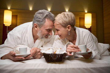 Front view of happy caucasian couple drinking tea or coffee on bed in hotel room
