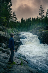 Boy Observing Thundering Waterfall in Dalarna Forest at Dusk. A young explorer stands on rocky terrain, captivated by the powerful rapids of a forest waterfall in Sweden, under a orange dramatic sky