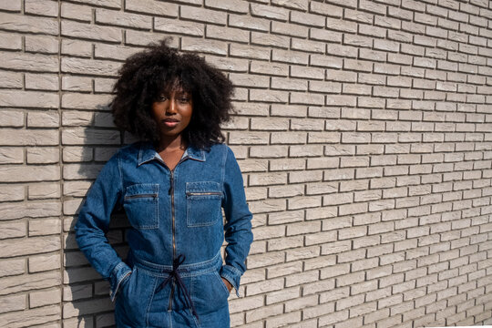 An African American woman with natural afro-textured hair stands confidently against a white brick wall backdrop. She sports a stylish full denim jumpsuit that cinches at the waist, complementing her