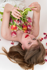 Obraz na płótnie Canvas Half naked romantic woman with spring tulip flowers lying on her chest and covering her, looking at camera. High angle view of beautiful blonde woman holding flowers, lying down on white background
