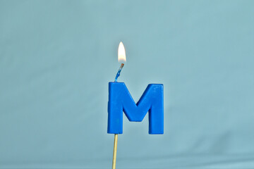 close up on a blue letter M birthday candle with fire on a white background.

