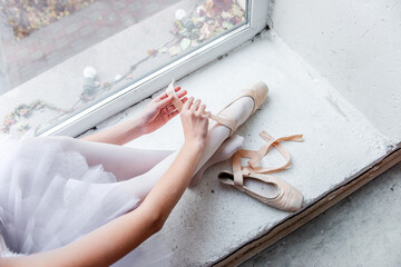 Overhead view captures a ballerina seated by a bright window, meticulously tying her pointe shoes...