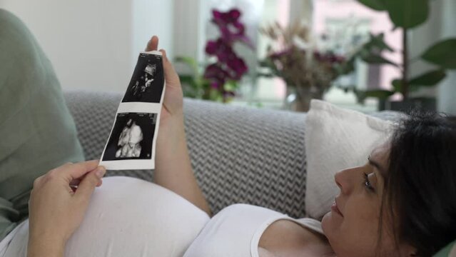 Pregnant woman looks at ultrasound image of her baby during late stage pregnancy laid on couch resting at home embracing maternal joy