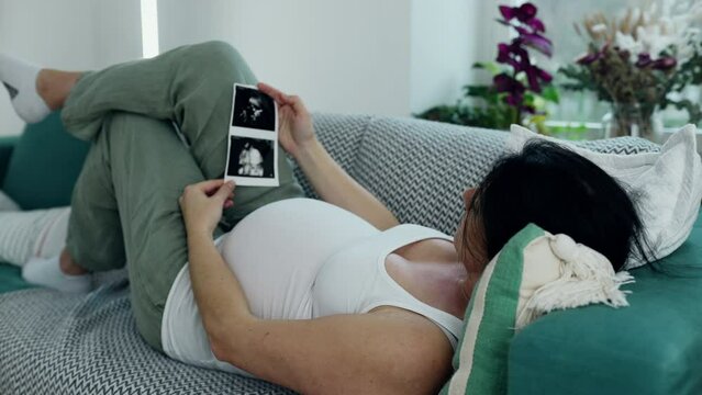 Pregnant woman laid on couch looking at ultrasound image of her baby during third trimester pregnancy. Expecting parent awaits her newborn