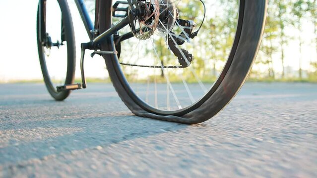 A man rolls a bicycle with a punctured wheel on the asphalt against the backdrop of the summer sun, close-up. Slow motion