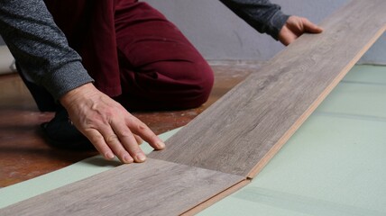 man joining two planks of beige textured laminate on the floor in the room, close-up, assembling wooden flooring on green soft mat during renovation