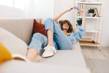 Smiling woman holding mobile phone, enjoying online games and reading messages on cozy couch in the living room.