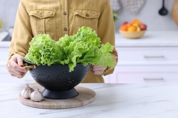 Woman holding black colander with lettuce at white marble table in kitchen, closeup. Space for text