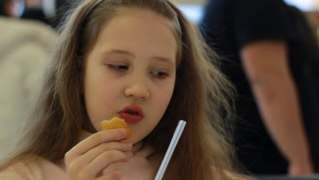 Closeup portrait of funny baby girl eats chicken nuggets and drinks a drink through a straw in the cafe. Child showing thumbs up and emotion of pleasure from eating. Fast food concept