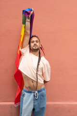 A man is holding a rainbow flag and wearing a pink shirt. He is standing in front of a red wall