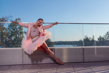 Sexy Slim Professional Caucasian Ballet Dancer in Pink Tutu Dress Posing in Standing Pose With Straight Legs On Glass Fence Outdoor.