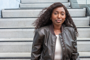An elegant Black woman sits comfortably on concrete stairs, her posture relaxed and confident. She wears a chic leather jacket over a white top, her hair flowing naturally. The urban setting and her - obrazy, fototapety, plakaty
