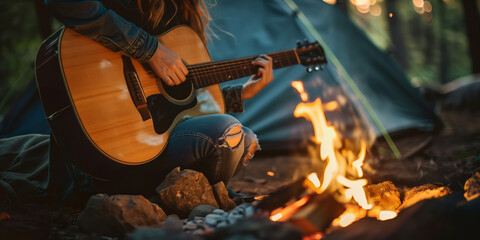 Cheerful young female musician playing a guitar by a bonfire at a campsite on summer night. - obrazy, fototapety, plakaty