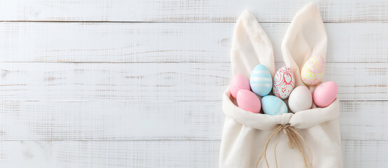 A cloth bag with bunny ears filled with colorful Easter eggs against a wooden backdrop. Holiday celebration and Easter concept. Festive minimalist banner with copy space.