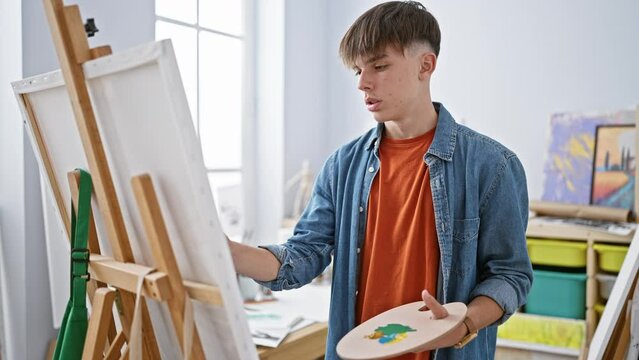 A young male artist in a studio attentively paints on a canvas, holding a palette in a creative environment.