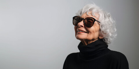 Portrait of an elderly woman wearing sunglasses against a gray wall on a sunny day.