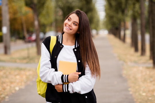 Photo of intelligent student with straight hairstyle dressed stylish jacket backpack on shoulder hug book go to school in autumn outdoors