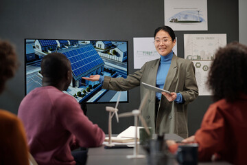 Happy young Asian businesswoman with tablet pointing at interactive board during presentation of...