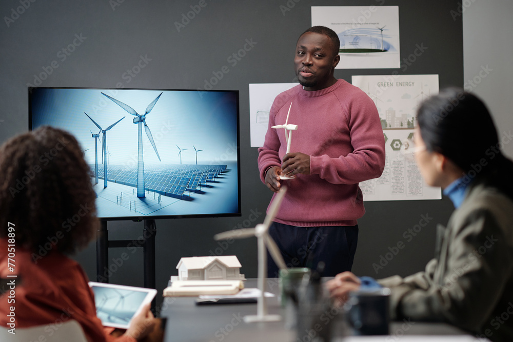 Poster young businessman with model of windmill in hands looking at female colleague while standing by inte