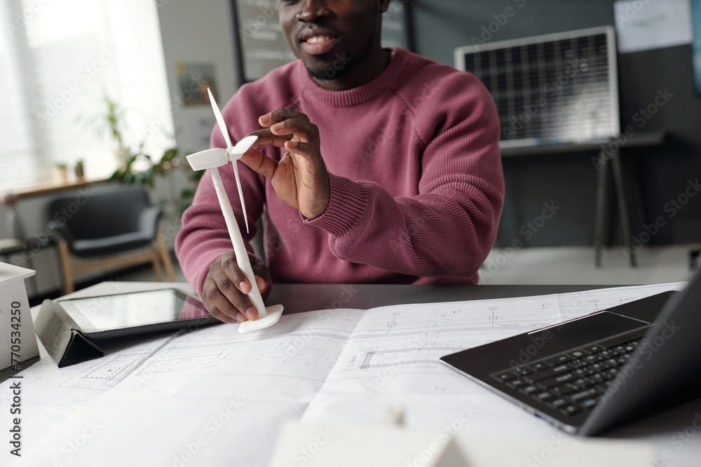 Poster Cropped shot of young modern architect rotating turbine of windmill model while sitting by desk with blueprint and mobile gadgets