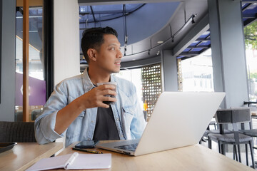 Asian man drinking a cup coffee with blue shirt using laptop and mobile phone in coffee shop, online freelance business