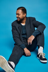 handsome african american man sitting with bent leg and looking to side on blue background