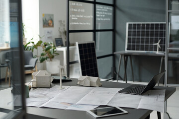 Blueprint with sketches surrounded by digital tablets, laptop and models of houses and windmill on workplace of modern architect in office