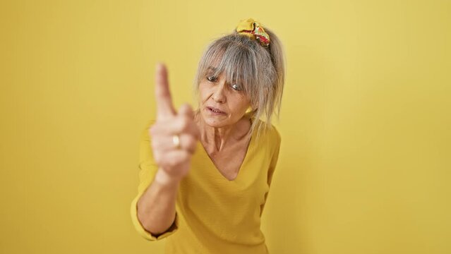 Angry middle-aged woman with grey ponytail furiously pointing at camera, blaming you! frustrated stance against isolated yellow background screams displeasure.