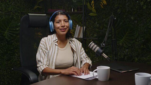 Portrait of young Latin female podcaster in headphones smiling and posing for camera at desk with mic, teacup and paper plan in recording studio