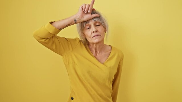 Middle-aged woman mocks and shows loser sign, teasing and laughing. grey hair in a ponytail, insulting gesture with fingers on forehead, isolated on a yellow background.